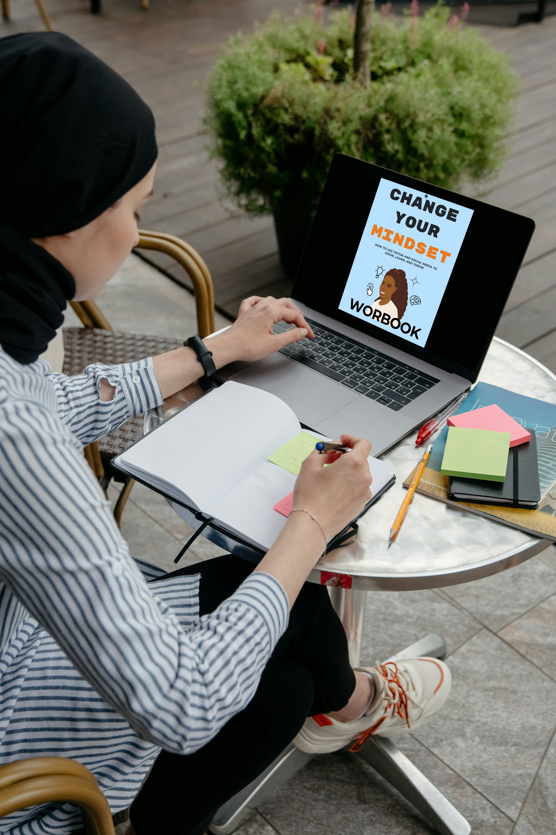Person using a laptop with a workbook titled 'Change Your Mindset' on a table outdoors.